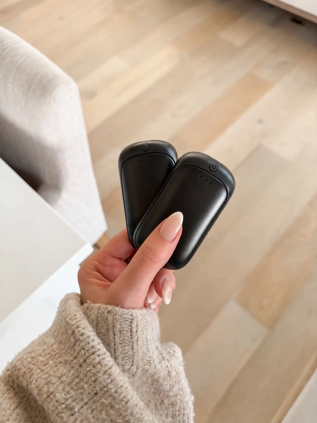 A hand in a beige sweater holding two black rechargeable hand warmers